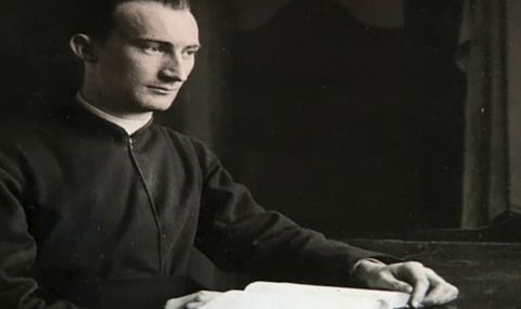 Mladi svećenik u crnoj reverendi sjedi za stolom nad otvorenom knjigom i gleda u stranu.